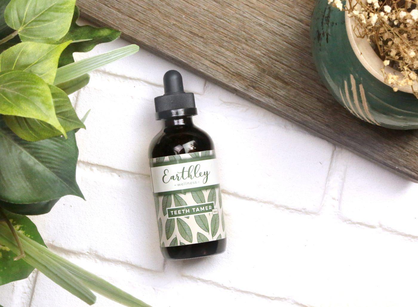 Earthley Teeth Tamer amber glass dropper bottle with black cap and dark green leaf pattern label laying on white brick surface with gray weathered wooden board teal green ceramic plate with dried babys breath flowers and green basil leaves
