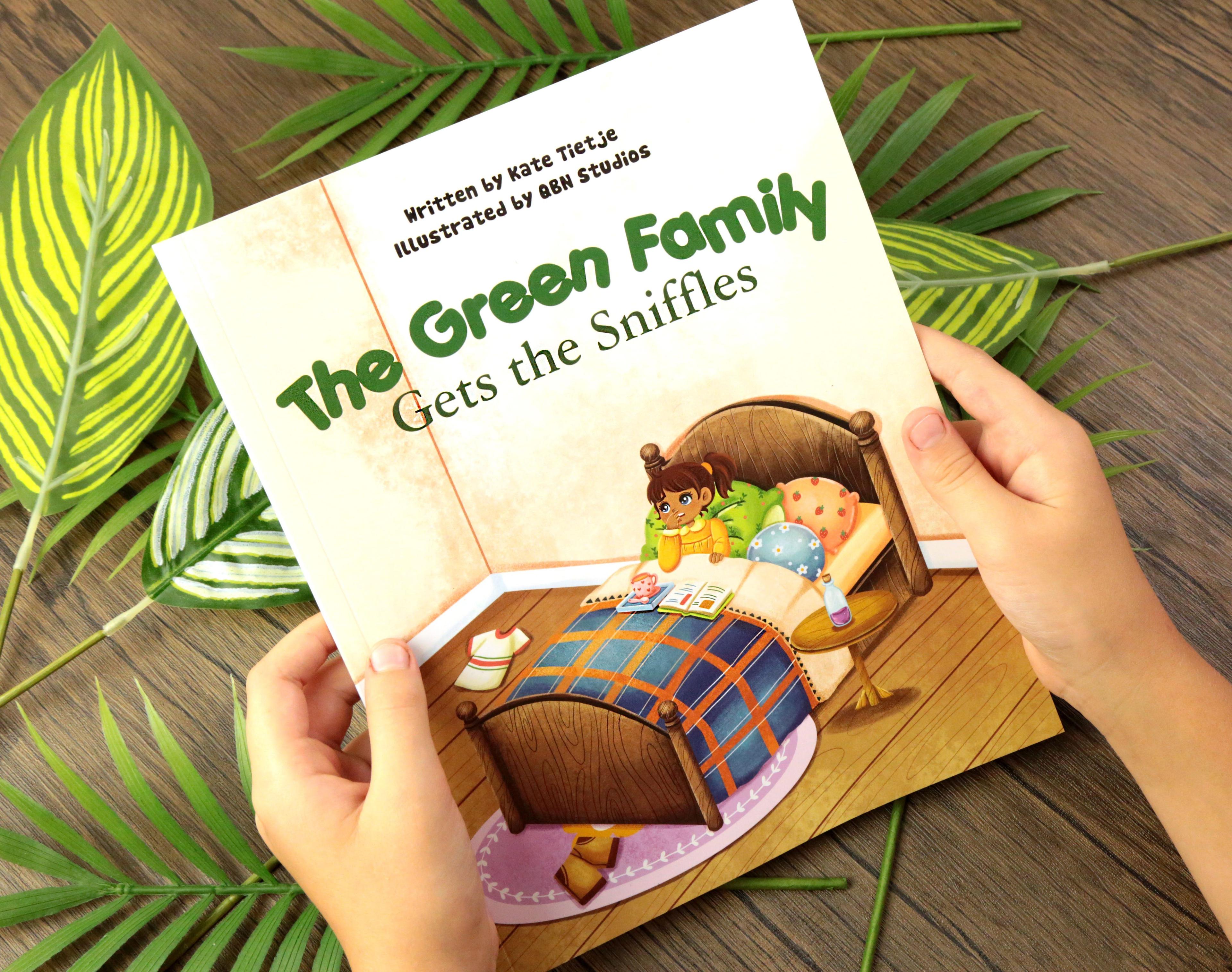 The Green Family Gets the Sniffles illustrated childrens book held by child hands showing cover with brown-haired girl in bed with blue orange plaid blanket on dark brown wooden surface with green tropical palm fronds and variegated white green leaves