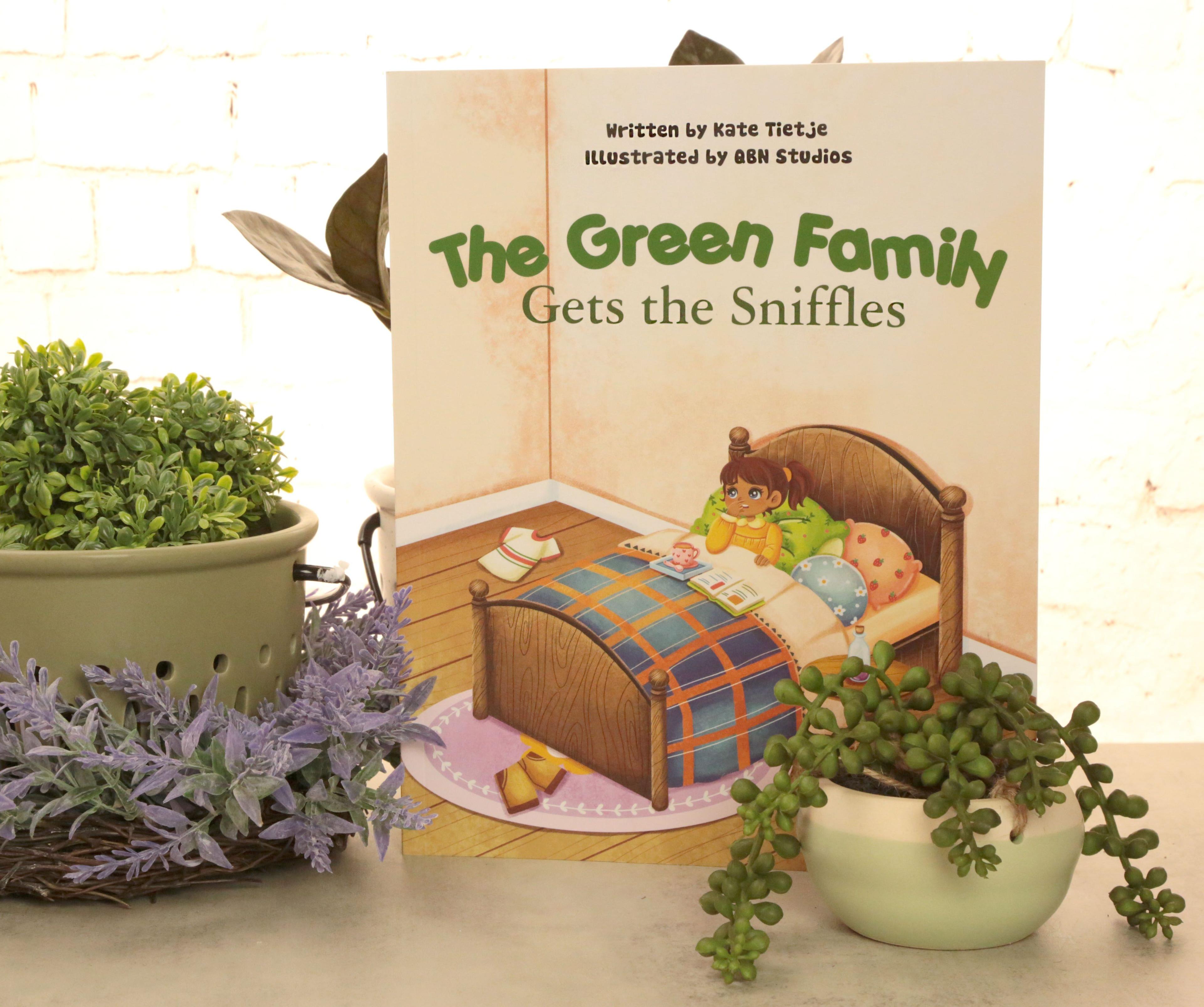 The Green Family Gets the Sniffles illustrated childrens book standing upright on tan surface with white brick wall background green plant in sage green colander purple lavender sprigs trailing green string of pearls succulent in cream ceramic pot