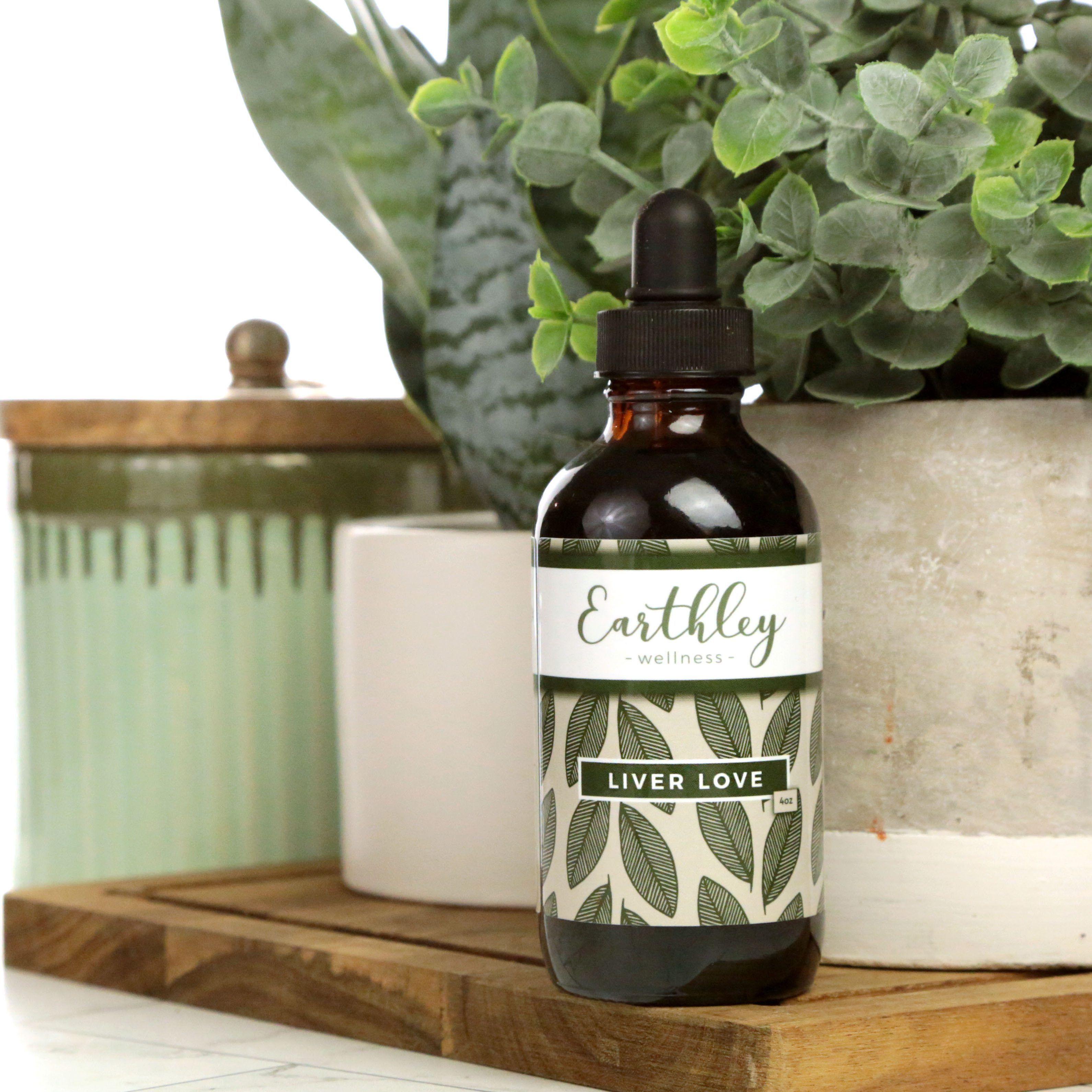 Earthley Liver Love 4oz amber glass dropper bottle with black cap and sage green olive leaf pattern label on wooden cutting board with mint green ceramic jar with wooden lid and green eucalyptus plant in cream concrete pot