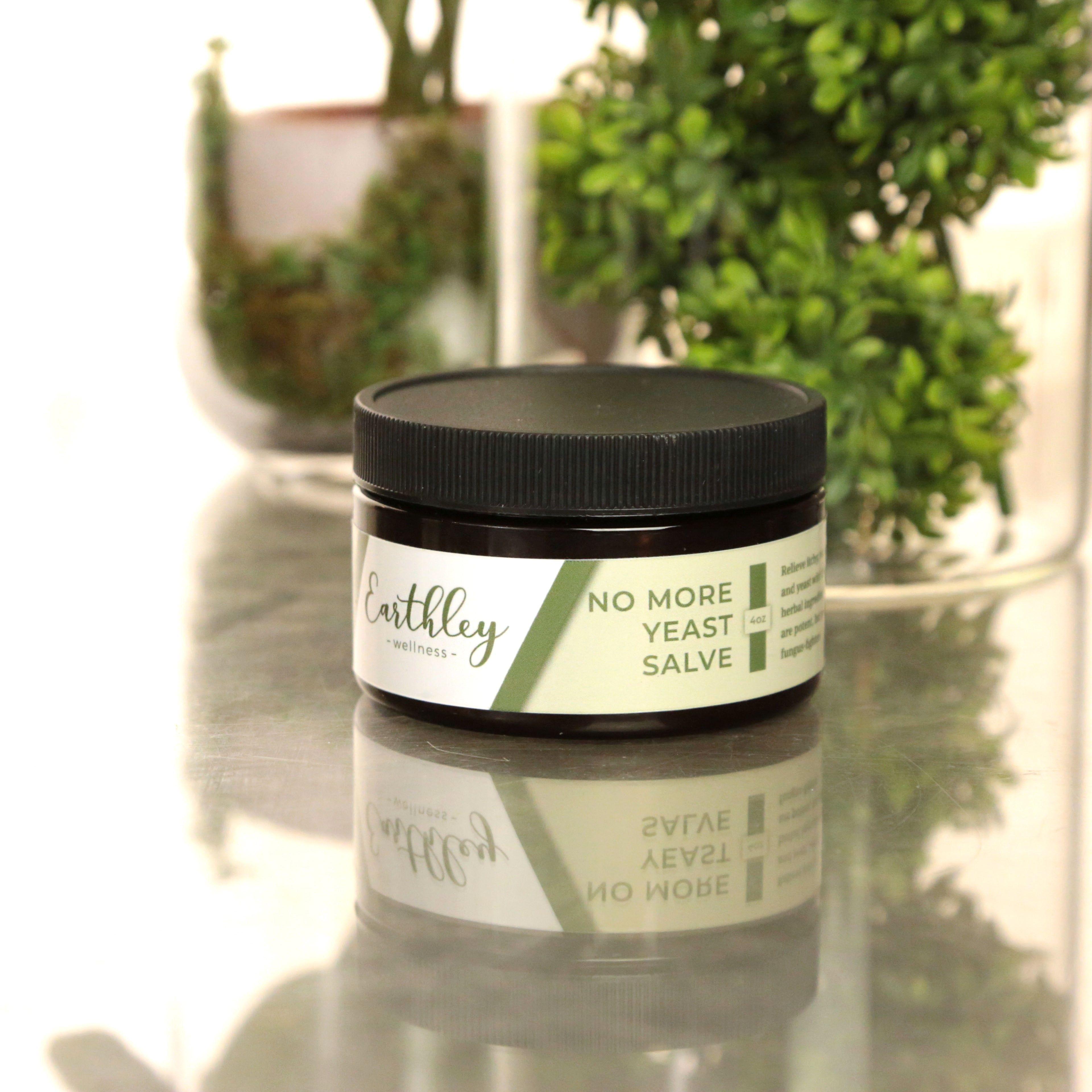 No More Yeast Salve 4oz product photo showing dark amber jar with black screw cap and cream white label with sage green stripe and text on reflective surface with green boxwood plants in background