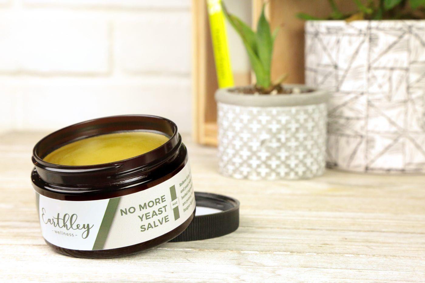 No More Yeast Salve 4oz open amber jar showing yellow balm with green stripe label, decorative pots with succulents in background