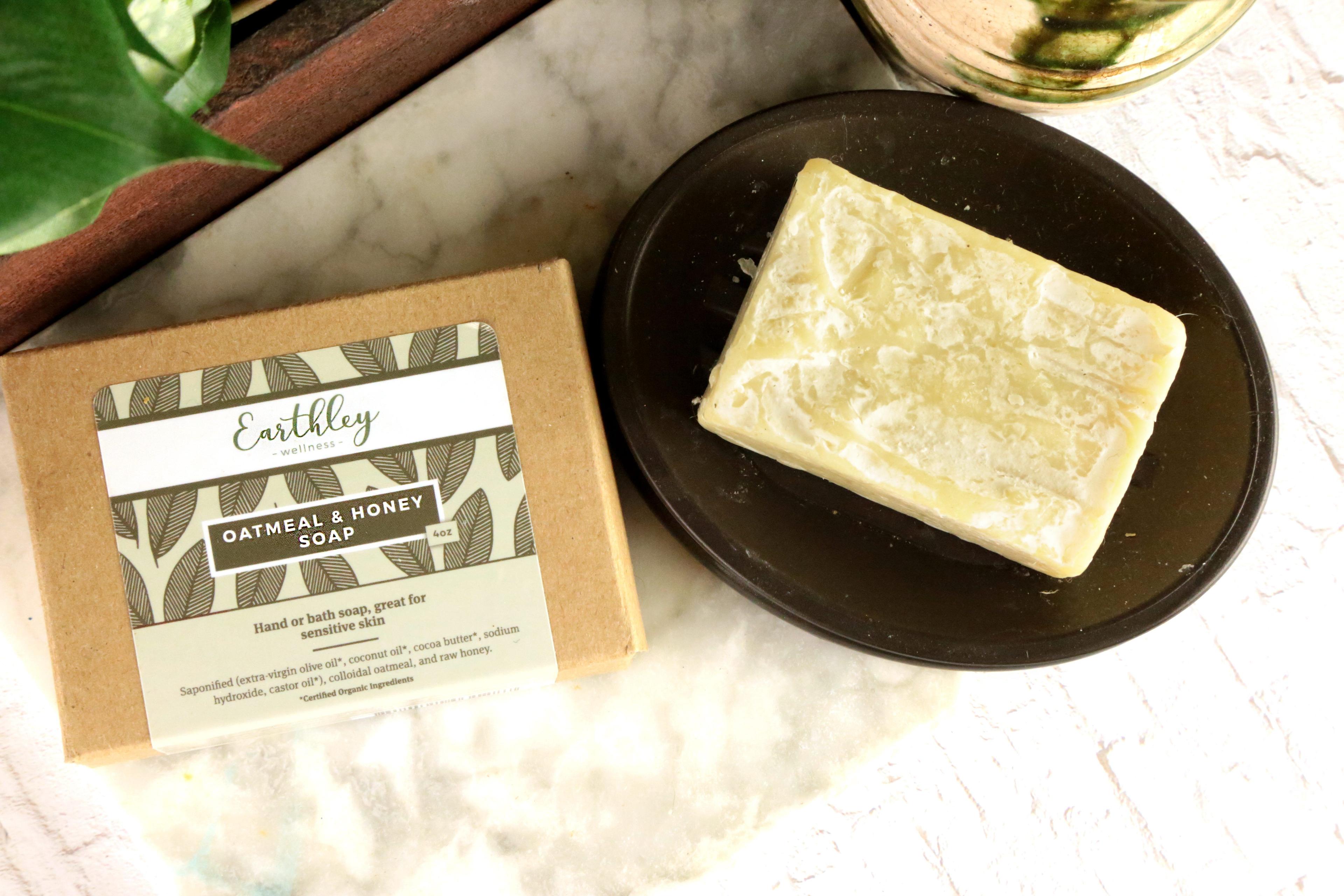 Oatmeal and Honey Soap kraft box with olive leaf label next to cream marbled soap bar on dark dish with greenery