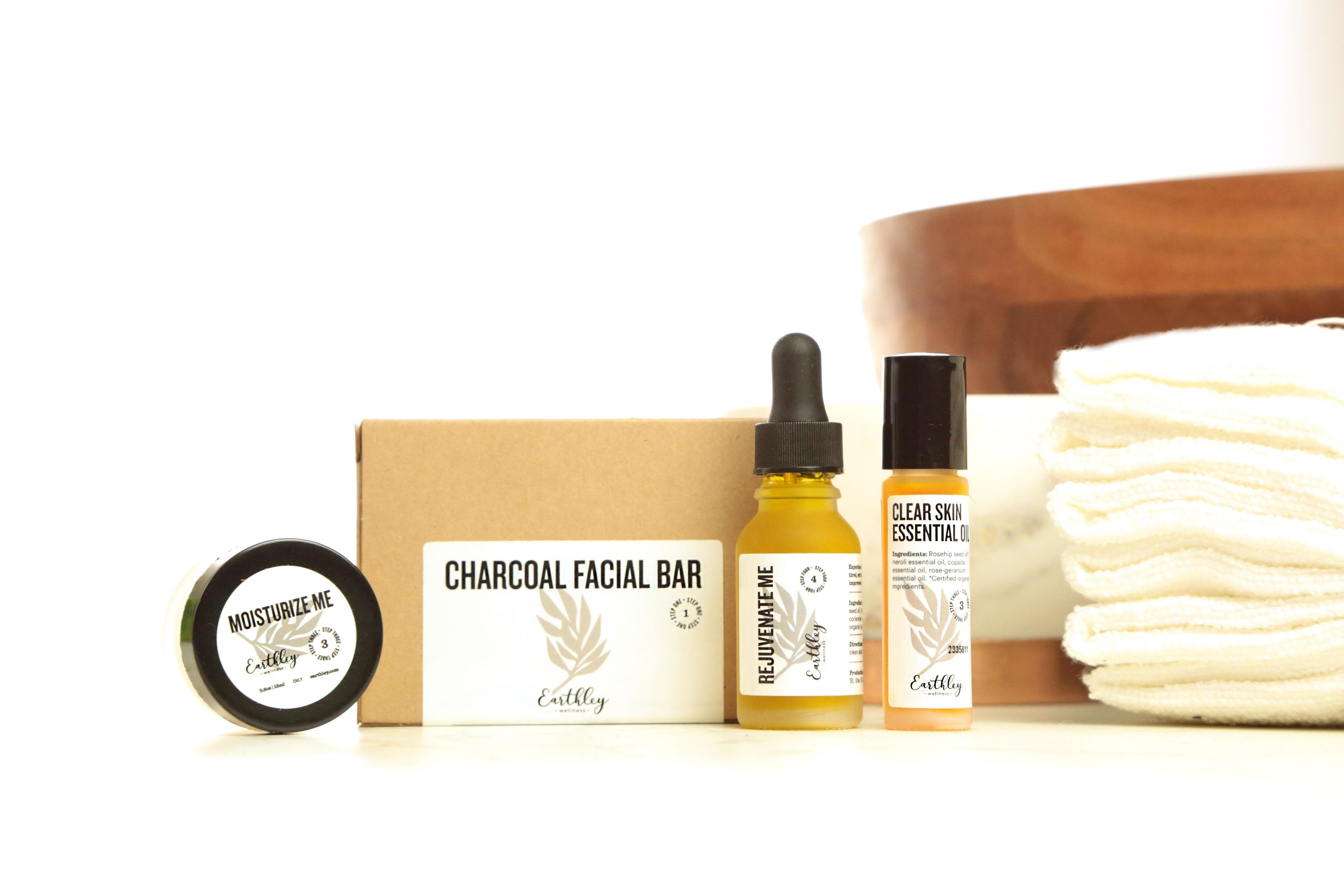 Earthley Oily Skin Bundle showing Moisturize Me jar, Charcoal Facial Bar box, Rejuvenate Me serum, and Clear Skin Essential Oil with white towels