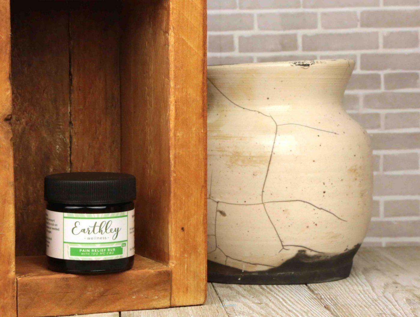 Pain Relief Rub with 100mg CBD in small amber jar with green stripe label in wooden shelf with rustic ceramic vase