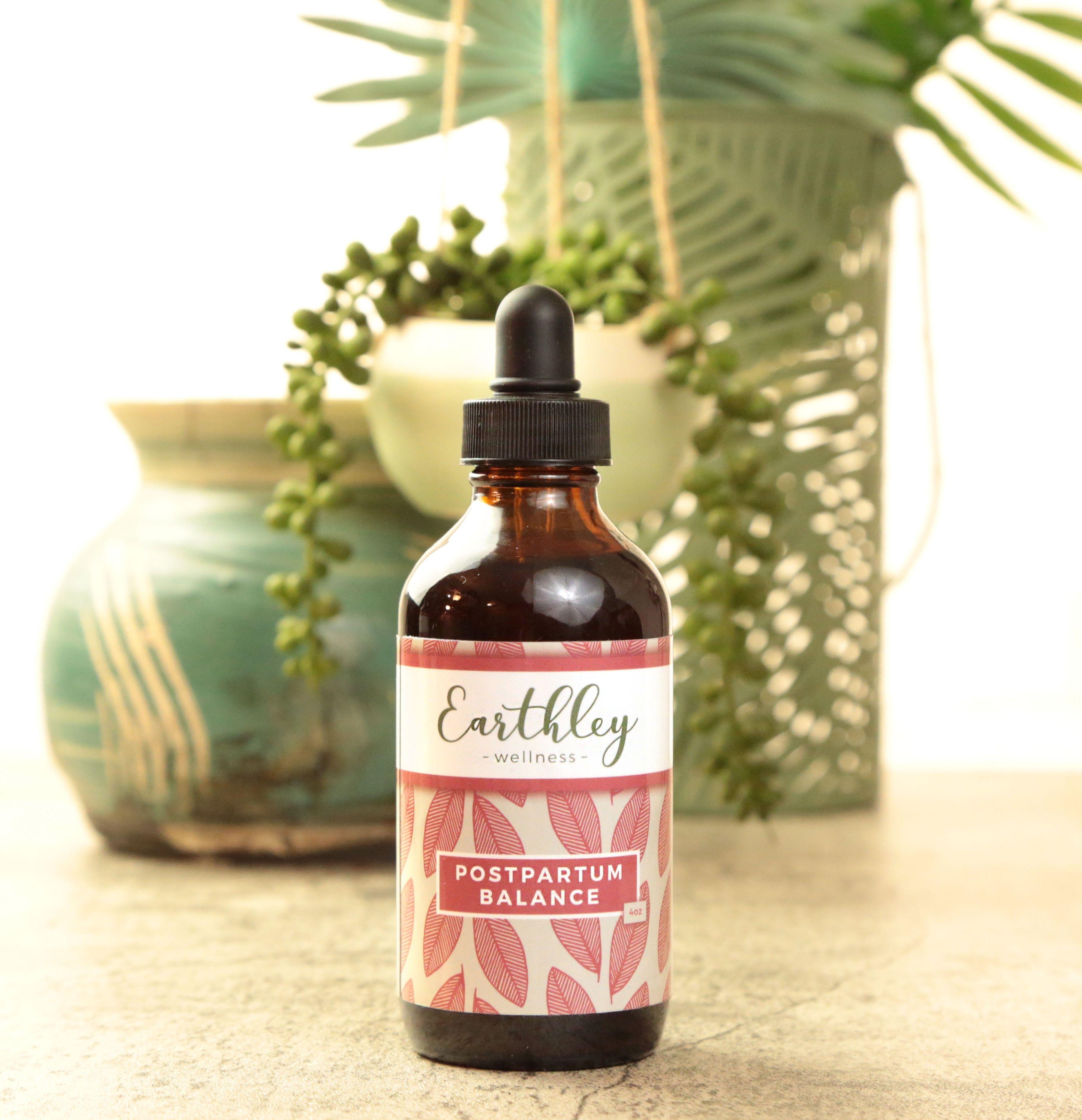 Postpartum Balance 4oz product photo showing amber glass dropper bottle with black cap and cream label with coral pink dusty rose leaf pattern on tan beige surface with teal turquoise ceramic vase with green plants in background