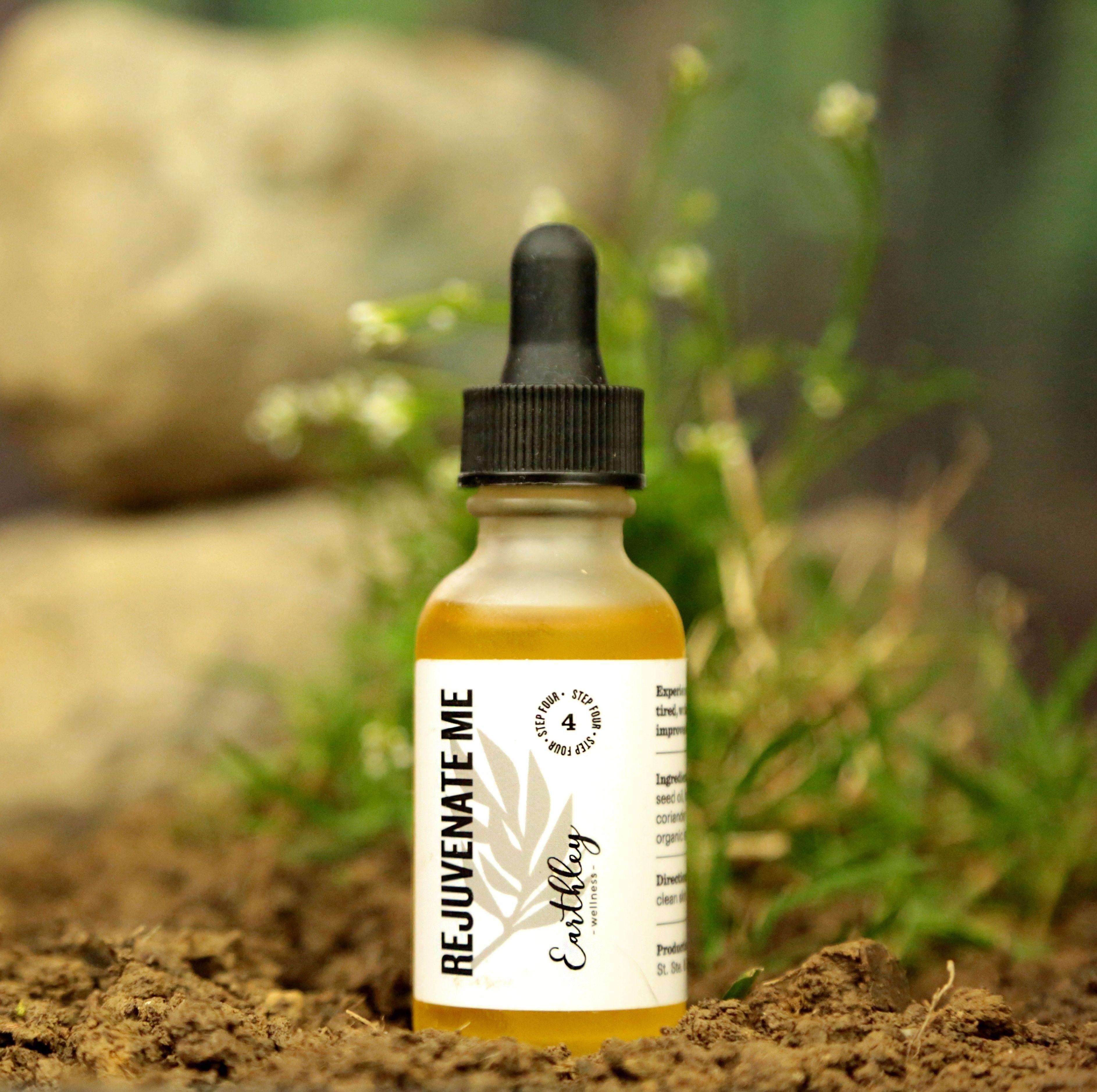 Earthley Rejuvenate Me Step 4 facial serum 1 oz amber dropper bottle with gray leaf label on soil ground with green plant sprouts and rock in background