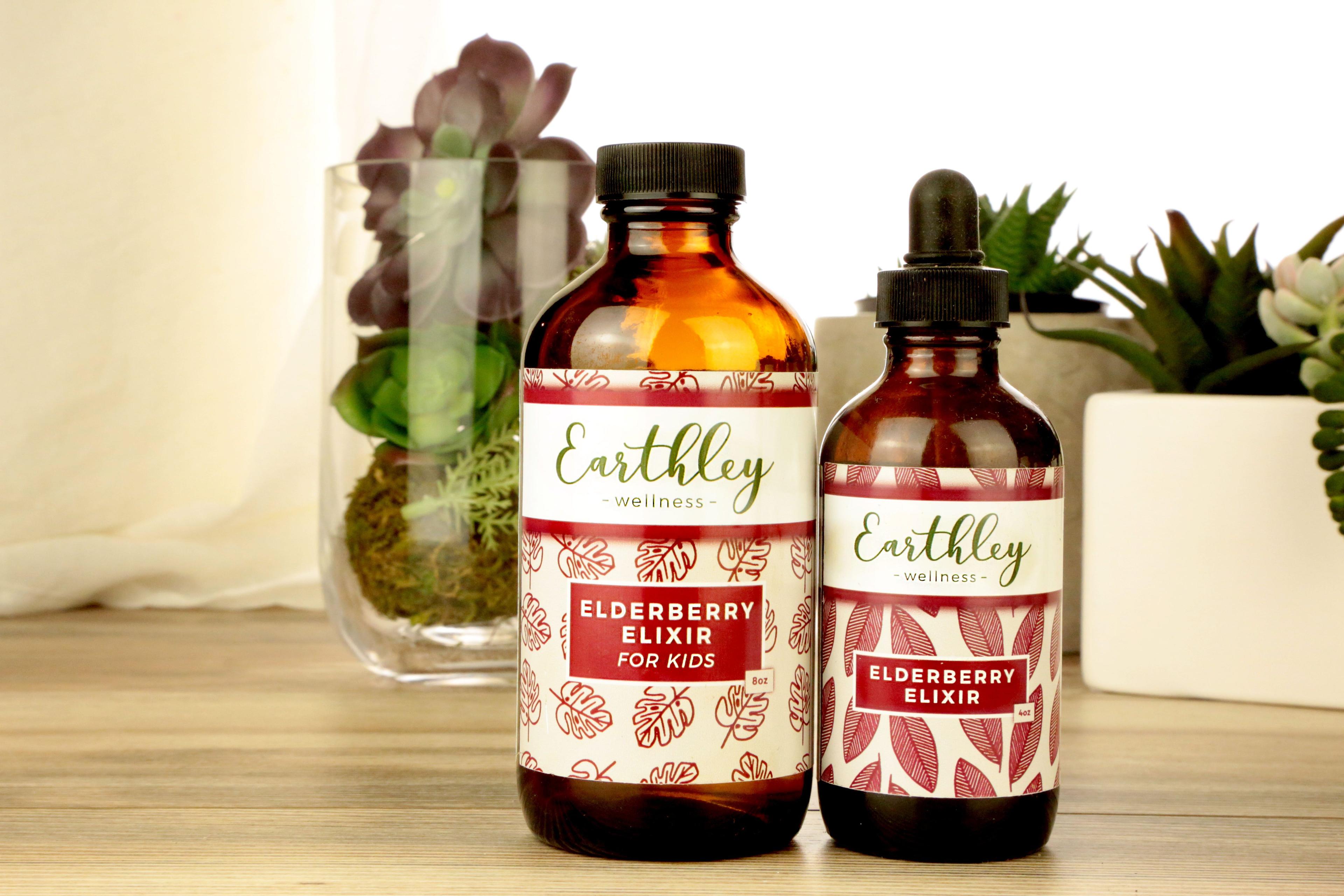 Earthley Elderberry Elixir For Kids 8oz amber glass bottle with screw cap and Elderberry Elixir 4oz amber dropper bottle both with burgundy red leaf labels with succulents in glass terrarium on wooden surface