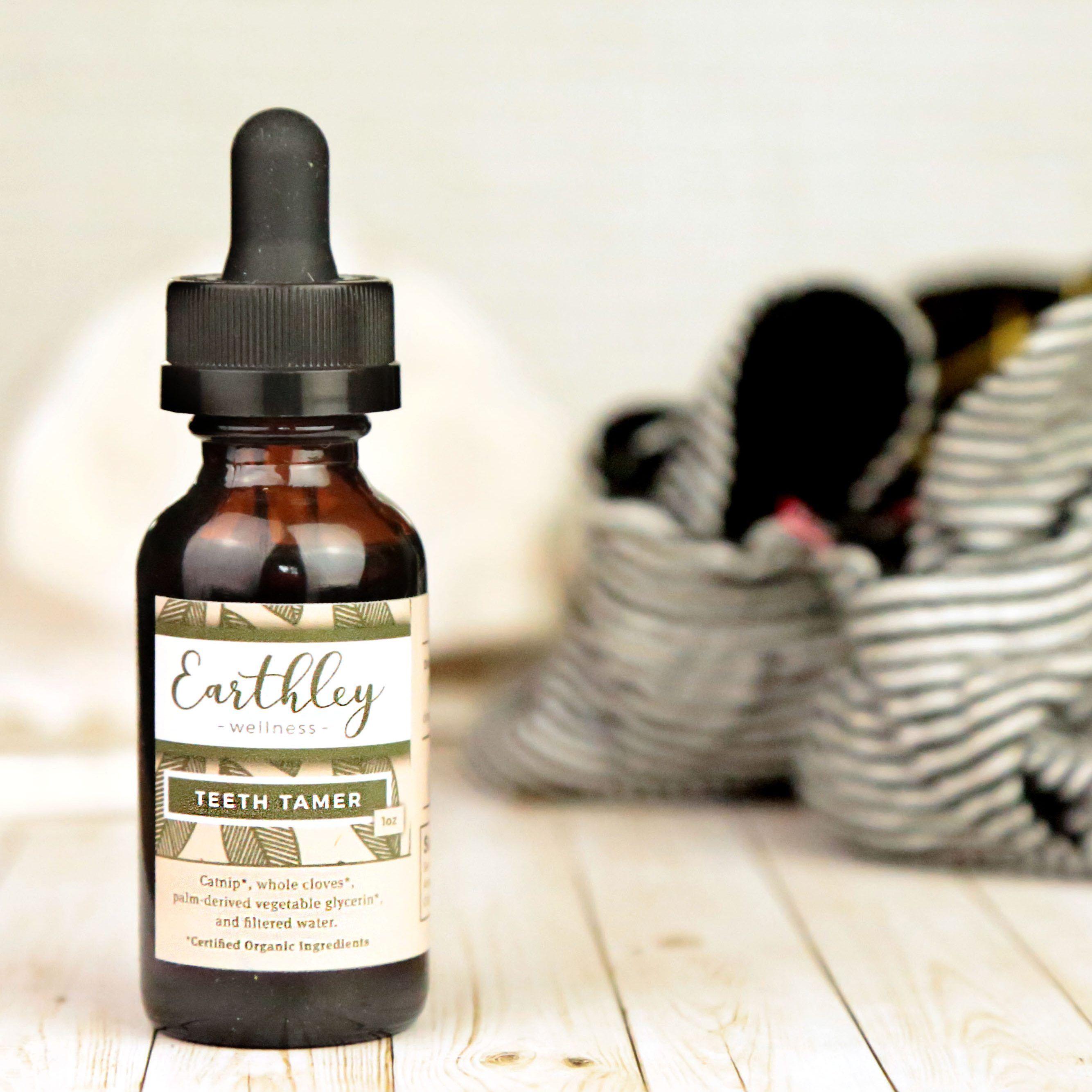 Earthley Teeth Tamer 1oz amber glass dropper bottle with black cap and olive green geometric pattern label on whitewashed wood surface with black and white striped baby shoes in soft focus background