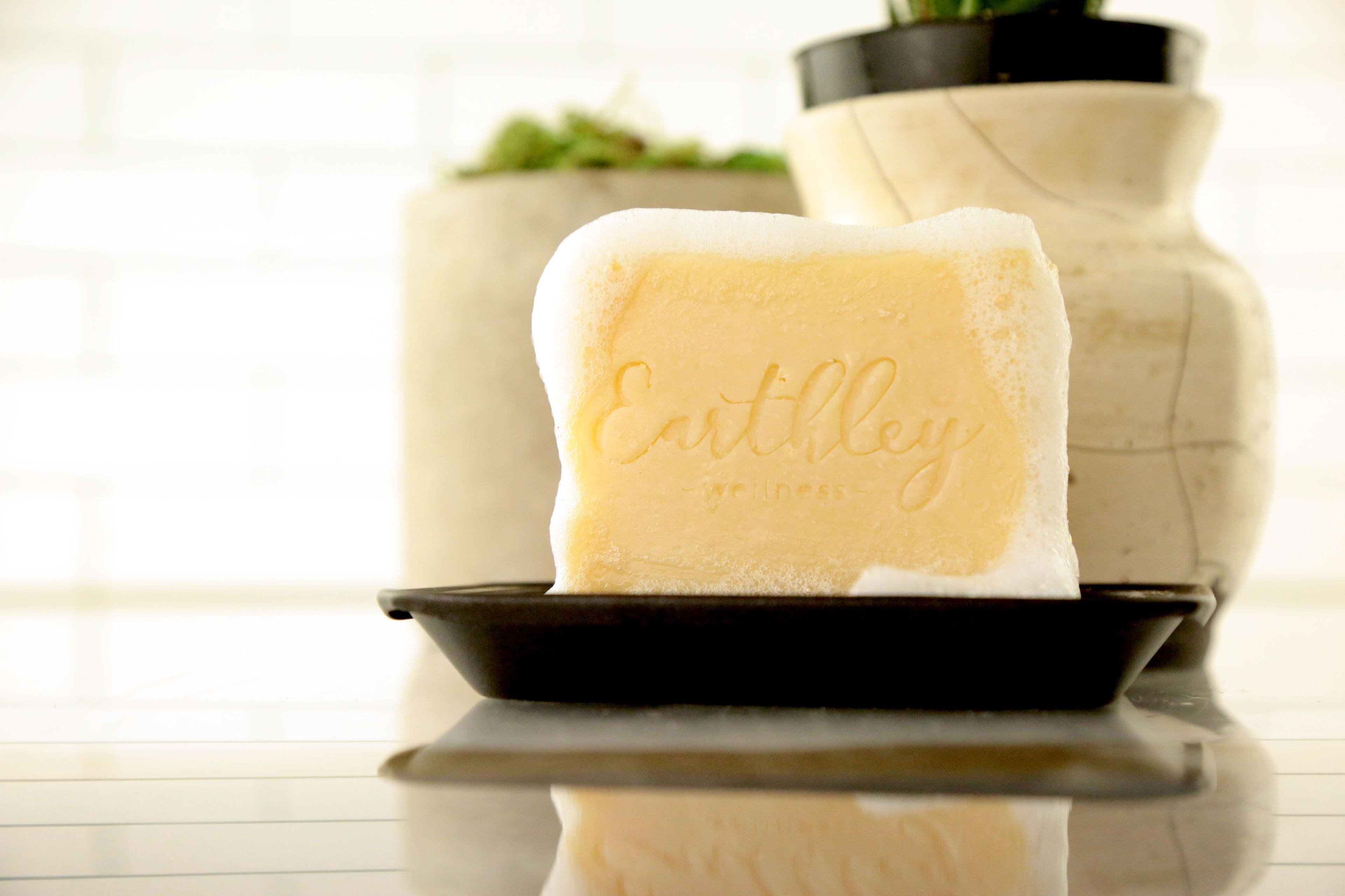 Earthley Shampoo Bar with embossed logo covered in foam suds on black soap dish with ceramic vases in background