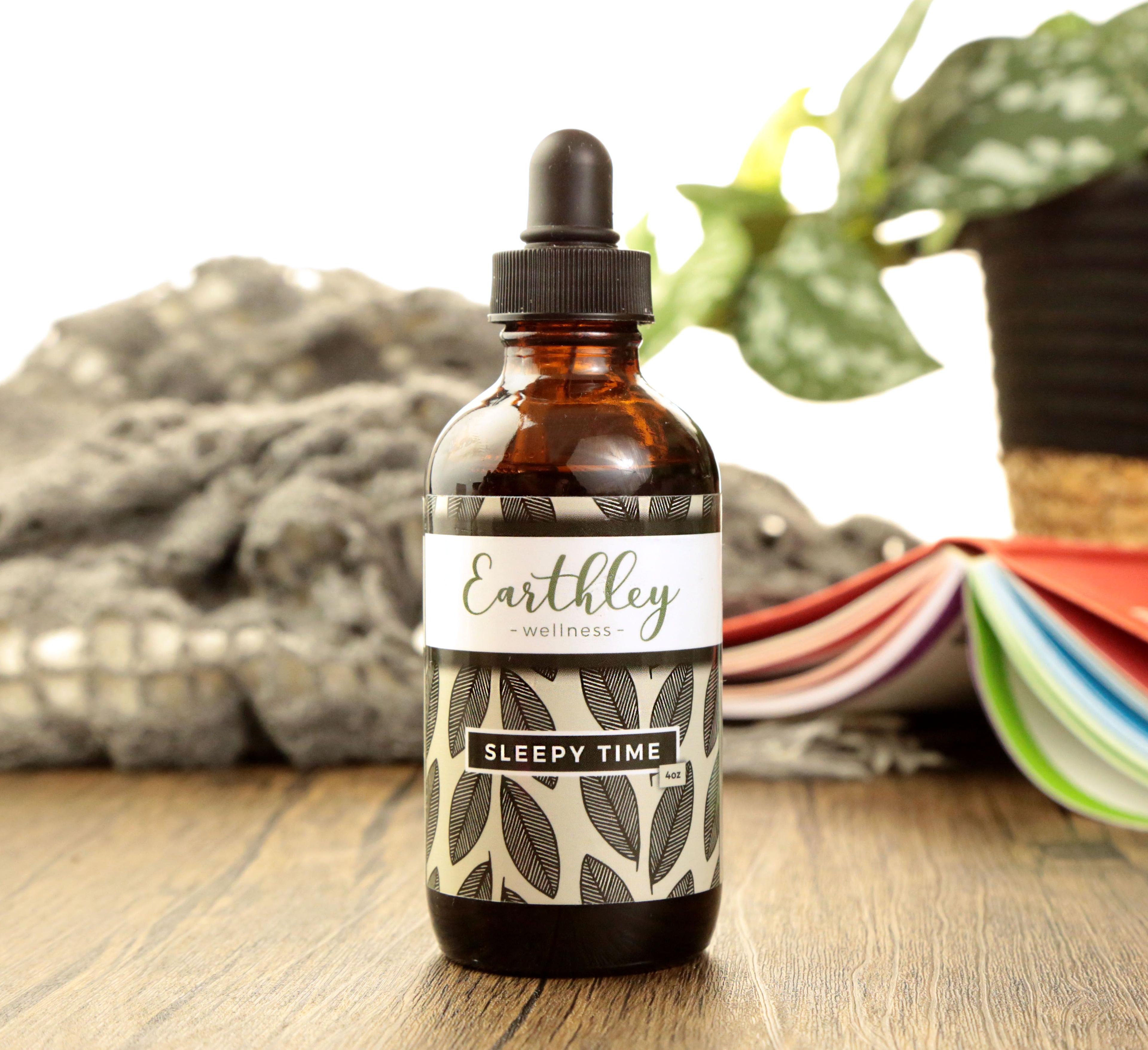 Earthley Sleepy Time herbal tincture 4oz amber dropper bottle with gray leaf pattern label on wood surface with cozy blanket colorful paper fan and potted plant in background