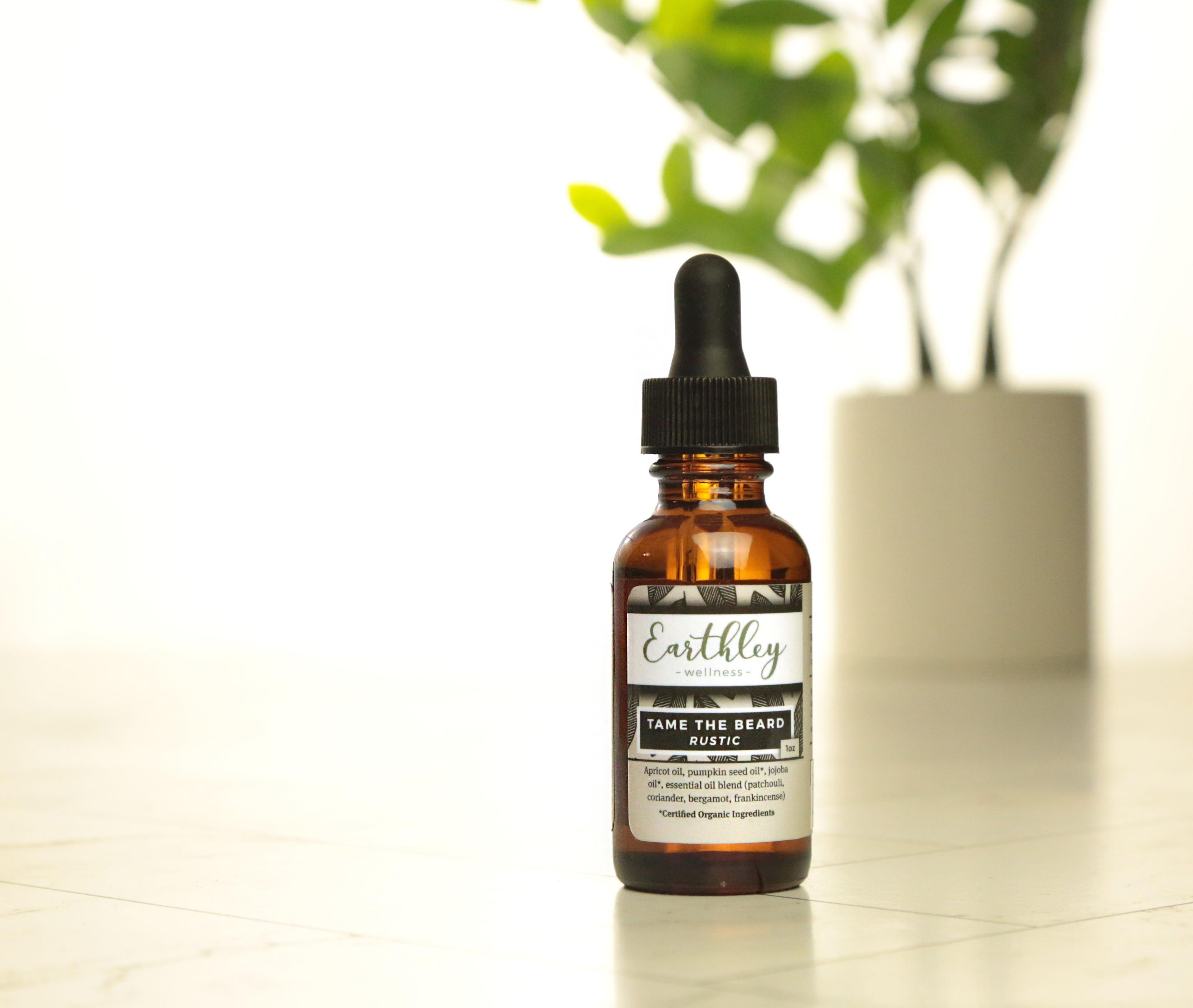 Earthley Tame the Beard Rustic scent 1oz amber dropper bottle with black and white geometric label listing apricot oil pumpkin seed oil jojoba oil and essential oils on marble surface with monstera plant