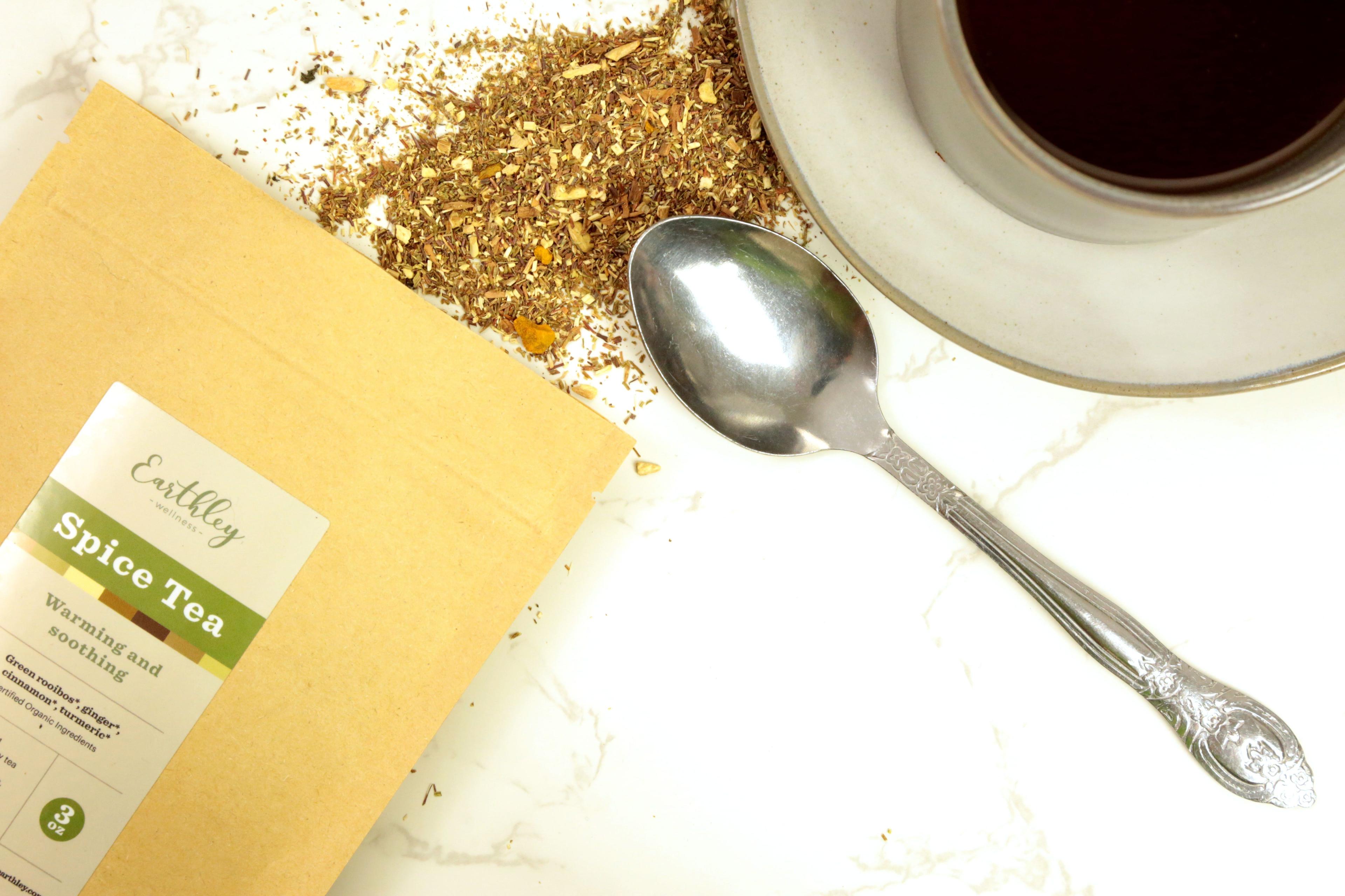Earthley Spice Tea 3oz tan kraft resealable pouch with sage green label on white marble with scattered brown tea blend silver ornate spoon and white ceramic cup and saucer
