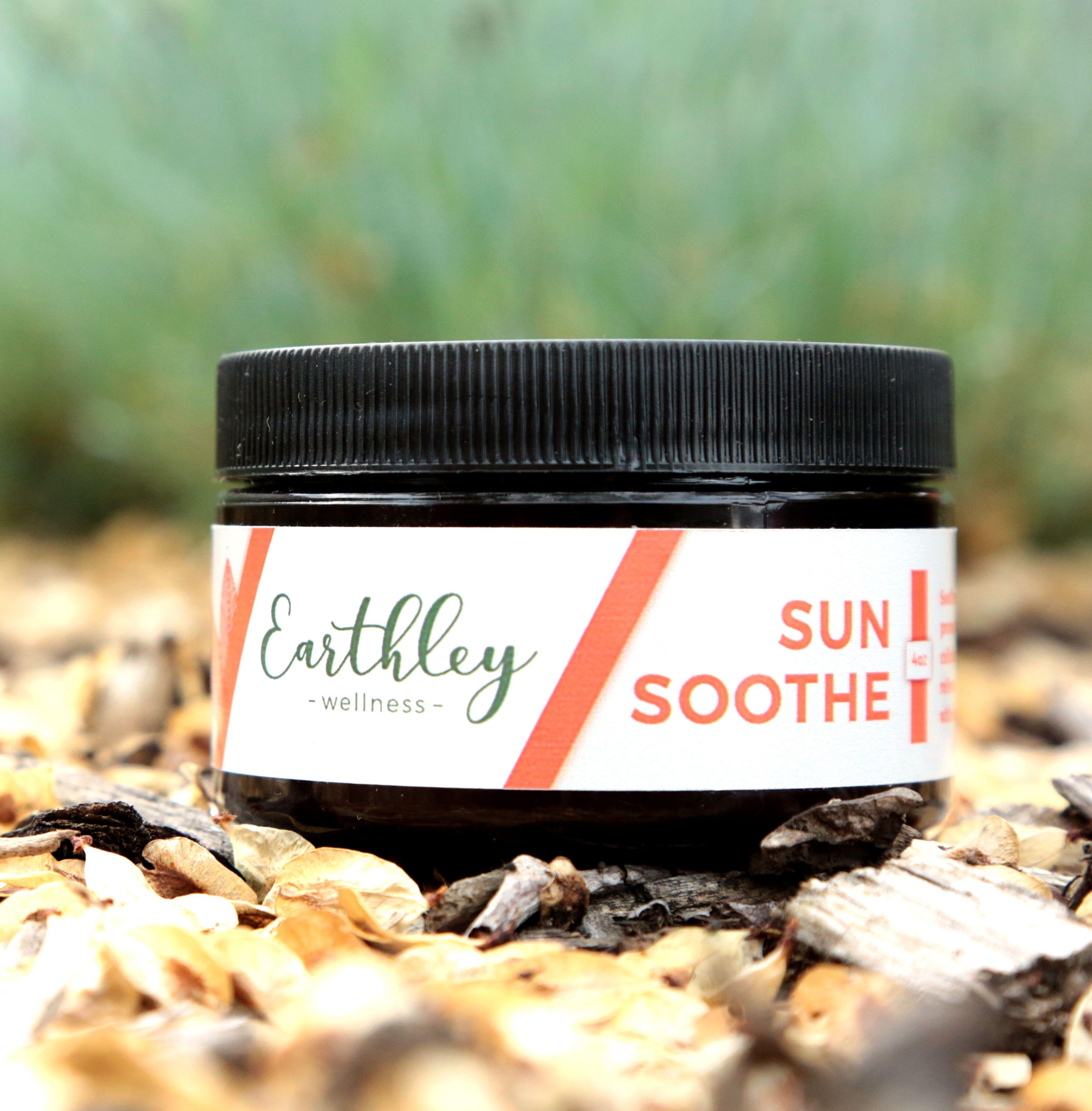 Sun Soothe product photo showing dark amber jar with black screw cap and white label with orange coral stripes on brown tan wood chips with green grass blurred in background