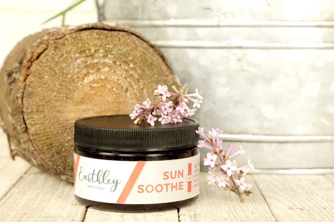 Earthley Sun Soothe 4oz amber glass jar with black lid and coral peach leaf pattern label on rustic white wooden surface with brown log slice pink lilac flowers and silver galvanized bucket