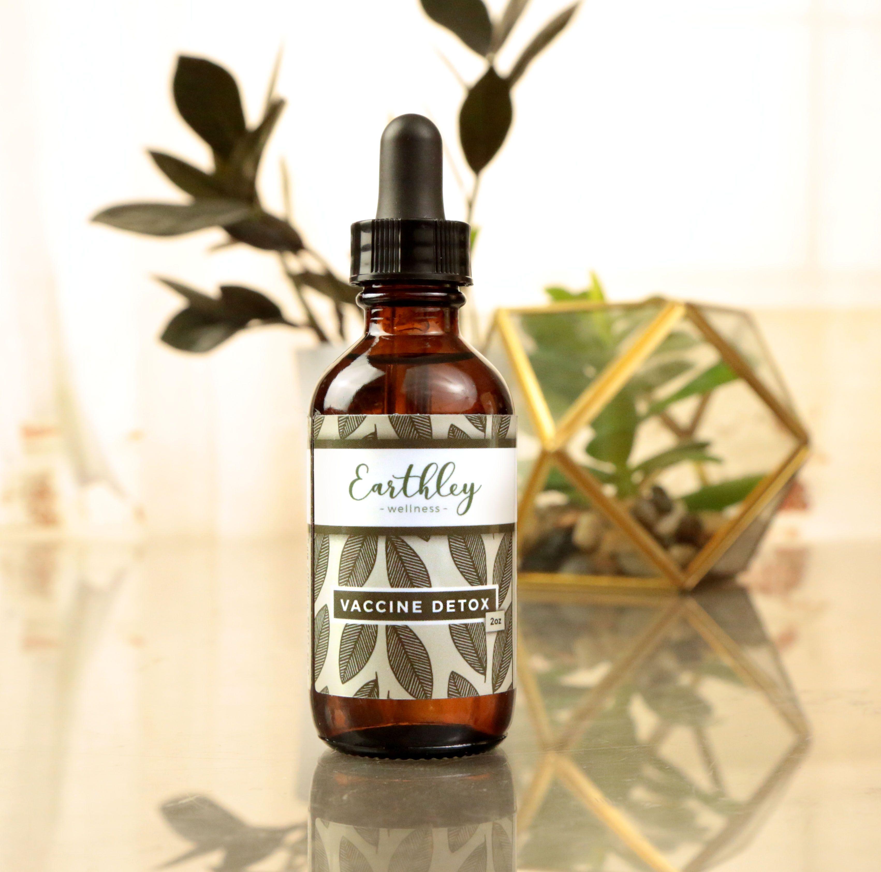 Vaccine Detox 2oz product photo showing amber glass dropper bottle with black cap and cream label with olive sage green leaf pattern on tan beige reflective surface with gold geometric terrarium containing green succulent in background