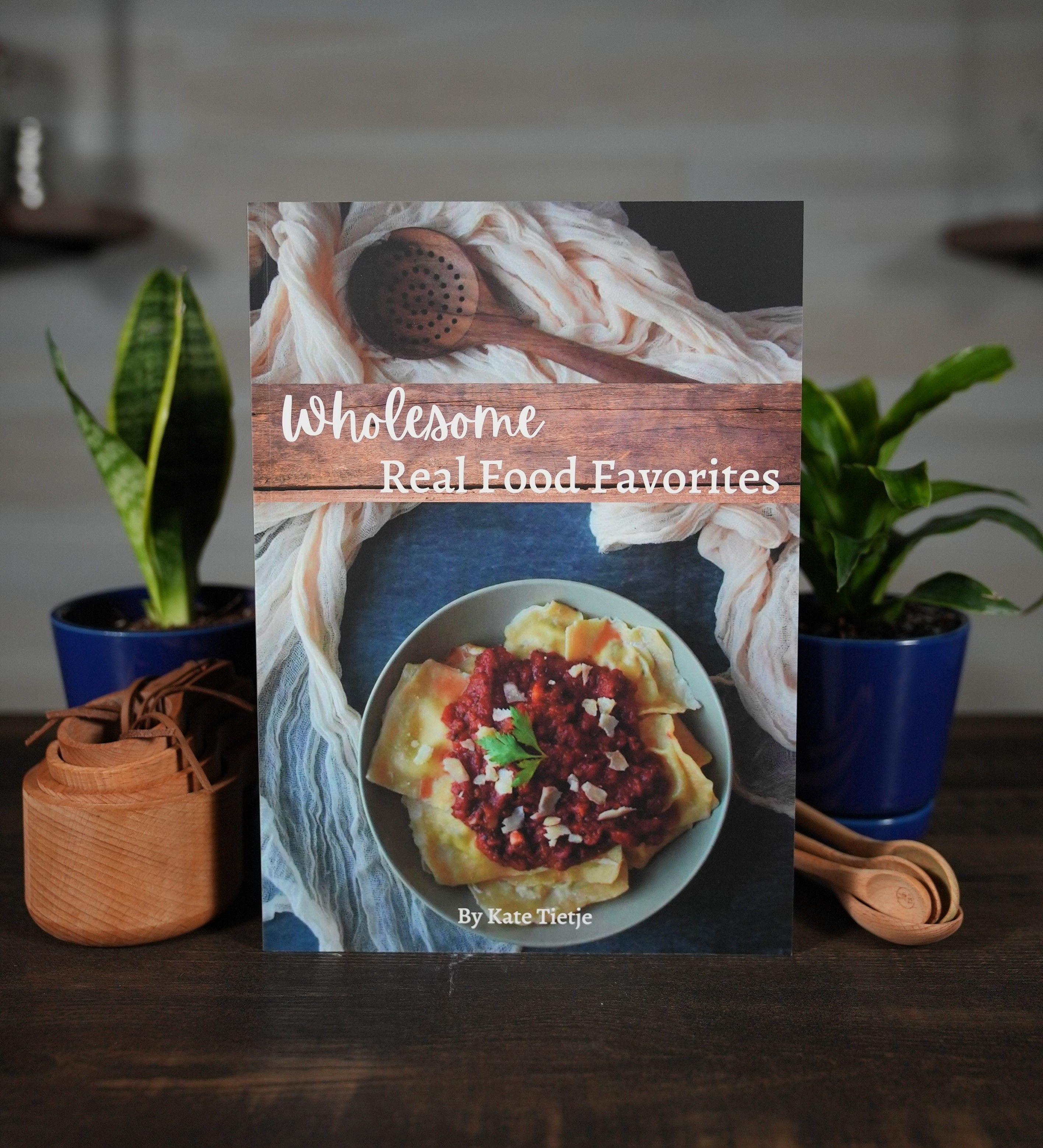 Earthley Wholesome Real Food Favorites Cookbook cover on dark brown wooden table with gray blue bowl of yellow pasta with red tomato sauce white cream towels brown wooden spoons and green snake plants in blue pots